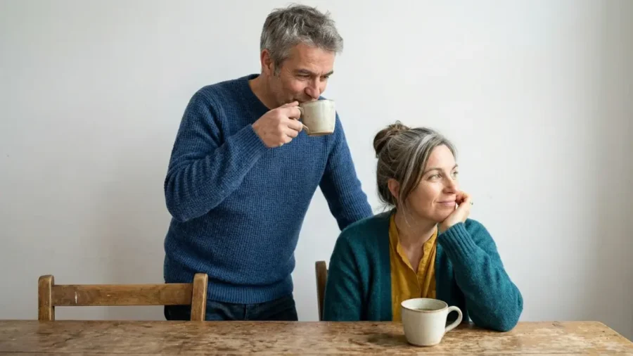 Klimakteriet för män. En man och en kvinna "i den där åldern" som dricker te. Hon sitter vid bordet och ser lite drömmande ut. Han står bakom henne och tittar på henne medan han smakar på sitt te.