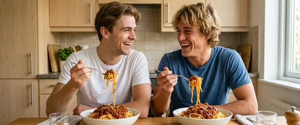 Två unga herrar som sitter och njuter av varsin stor tallrik spagetti i ett kök.