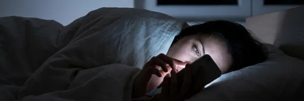 En kvinna ligger i sängen och fipplar med sin telefon, mitt i natten